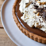 Chocolate Cinnamon Espresso Tart garnished with fresh whipped cream shaved chocolate and crushed peanuts on a white plate and wood background