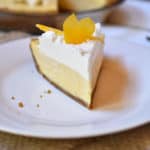 A slice of Lemon Cream Pie on a white plate with the remaining whole pie in the background