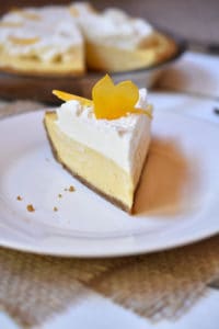 A slice of Lemon Cream Pie on a white plate with the remaining whole pie in the background