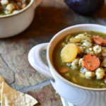 A bowl of Verde Pozole in a white bowl with hominy garnish, chips, and an avocado on a slate background