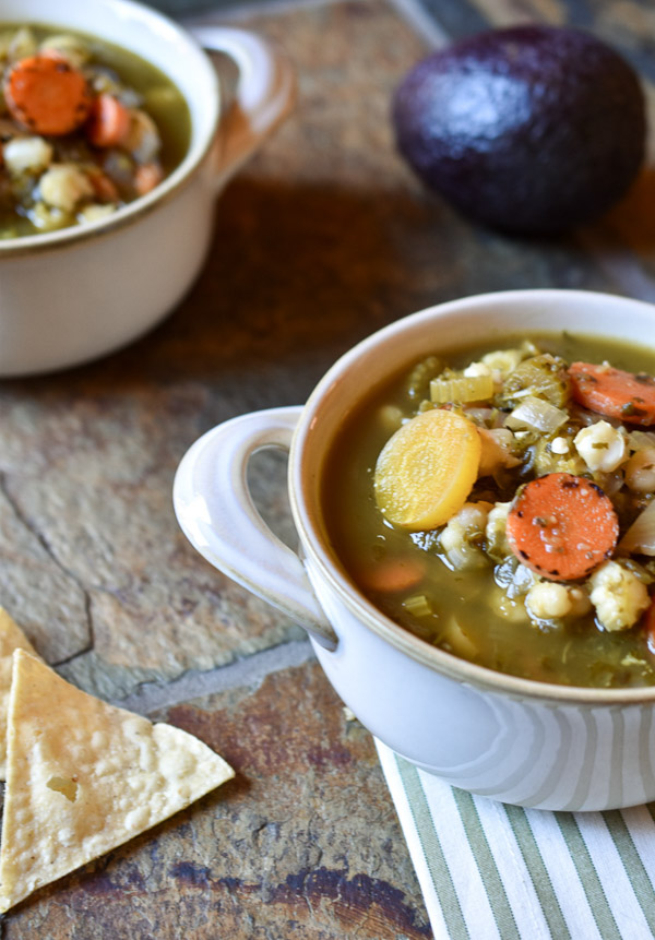 A bowl of Verde Pozole in a white bowl with hominy garnish, chips, and an avocado on a slate background