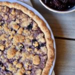 A spiced cherry berry pie on a grey and white napkin with a bowl full of berries to the upper right