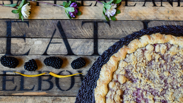 A blackberry orange mint pie on a wooden backdrop with the word "eat" garnished with whole blackberries, orange zest, and flowers