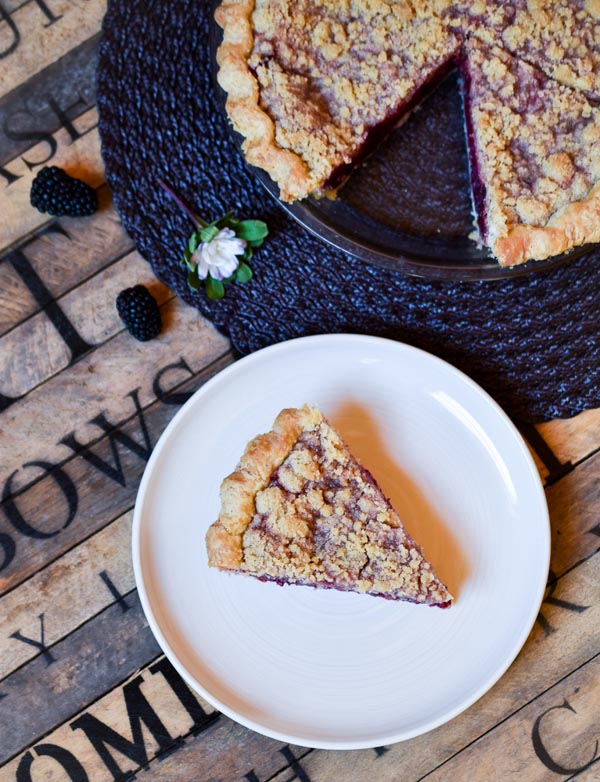 slice of blackberry orange mint pie with the whole pie in the background garnished with blackberries, and flowers