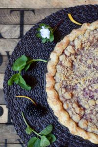A blackberry orange mint pie on a placemat garnished with orange zest, mint leaves, and flowers