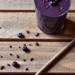 A blueberry power greens smoothie on a wood background with a bamboo straw and blueberry and cocoa nib garnish