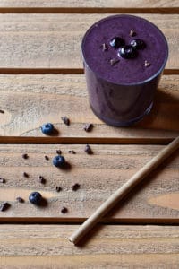 A blueberry power greens smoothie on a wood background with a bamboo straw and blueberry and cocoa nib garnish