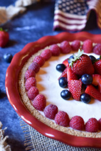 A whole cream cheese pie with strawberry, raspberry, and blueberry garnish and American flag in the background.