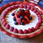 A whole cream cheese pie with strawberry, raspberry, and blueberry garnish and American flag in the background.