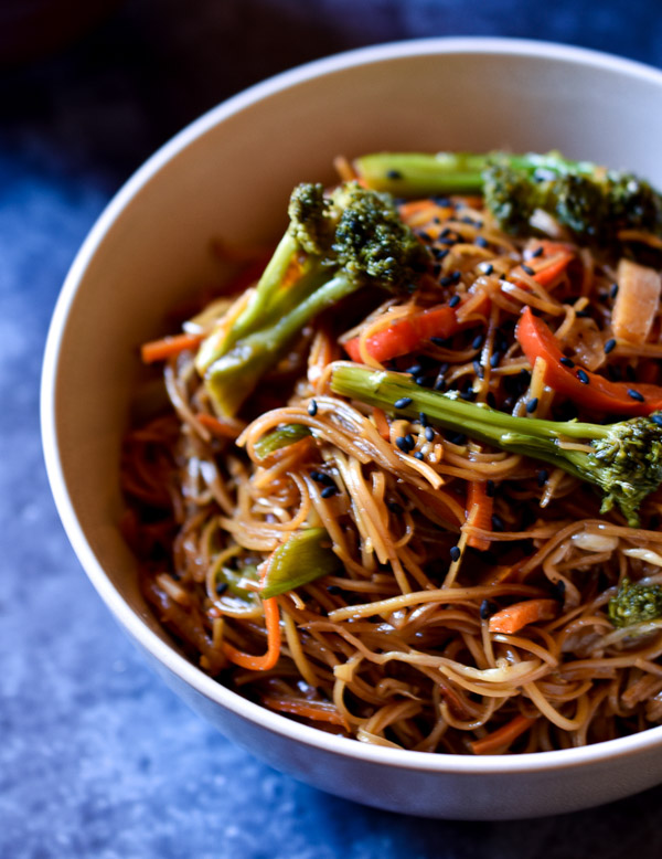 Vegetable chow mein in a white bowl with a dried flower to the left and a cup of tea to the upper right