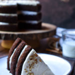 A slice of Fall Frosted Chocolate Cake with the rest of the cake and a cup of milk in the background.