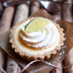 A key lime tarts garnished with fresh whipped cream dusted with powdered sugar, lime, and a fork and spoon in the background