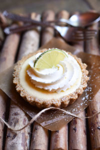 A key lime tarts garnished with fresh whipped cream dusted with powdered sugar, lime, and a fork and spoon in the background