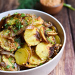 A bowl of roasted potato salad garnished with fresh dill and pickled mustard seeds in the background.