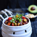 A bowl of vegetable chili in a mummy bowl with a skeleton hand, cheese, and avocado in the background