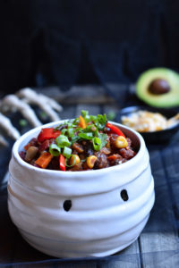 A bowl of vegetable chili in a mummy bowl with a skeleton hand, cheese, and avocado in the background