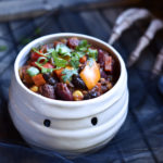 A bowl of vegetable chili in a mummy bowl with a skeleton hand in the background