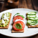 Three flatbread crackers: one topped with smashed avocado and feta, two with mozzarella, tomato and basil, three with cream cheese, cucumber, and dill.