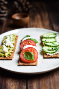 Three flatbread crackers: one topped with smashed avocado and feta, two with mozzarella, tomato and basil, three with cream cheese, cucumber, and dill.