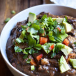 A bowl of Ancho Black Bean Soup garnished with avocado, cilantro , and red bell pepper.