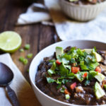 A bowl of Ancho Black Bean Soup garnished with avocado, cilantro , and red bell pepper. Another bowl is in the background along with a spoon and lime half.