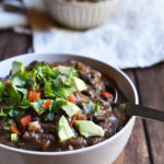 A bowl of Ancho Black Bean Soup garnished with avocado, cilantro, and red bell pepper.