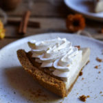 A slice of Autumn Spiced Cream Pie with another piece, cinnamon sticks, pecans, and burnt orange flowers in the background