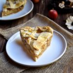 A slice of Brandy Apple Pie with another, apples, dried flowers, and cinnamon sticks in the background.