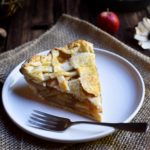 A slice of Brandy Apple Pie accompanied by a fork with apples, dried flowers, and cinnamon sticks in the background.