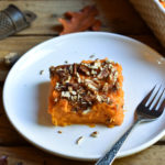 A helping of Pecan Yam Casserole on a plate with a fork. A background of fall leaves and the remainder of the casserole.