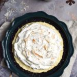 A Coconut Cream Pie in a cookie crust topped with fresh whipped cream and toasted coconut with branch twigs and coconut and metal snow flakes in the background.