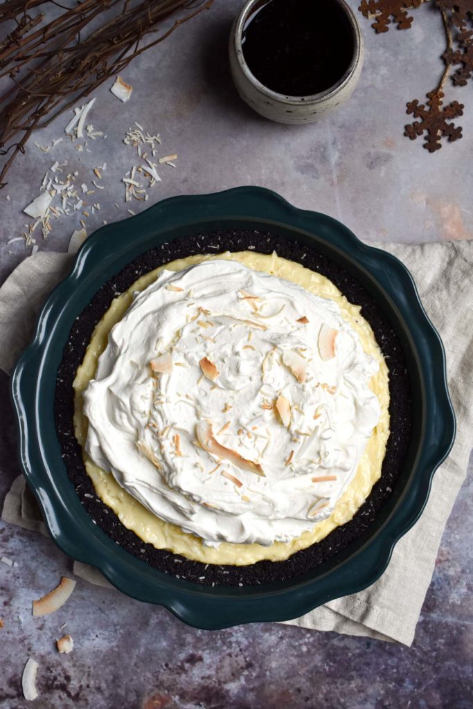 A Coconut Cream Pie in a cookie crust topped with fresh whipped cream and toasted coconut with branch twigs and coconut and metal snow flakes in the background.