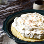 A Coconut Cream Pie in a cookie crust with branch twigs, coconut flakes, and a cup of coffee in the background.