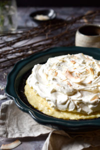 A Coconut Cream Pie in a cookie crust with branch twigs, coconut flakes, and a cup of coffee in the background.