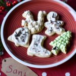 An assortment of frosted sugar cookies (bell, boot, tree, squirrel, gingerbread man) on a red and white plate with a tag and handwritten "Santa" on a red and white polka dot runner