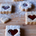 Heart and flower shaped powdered sugar dusted Linzer Cookies on a wood cutting board.