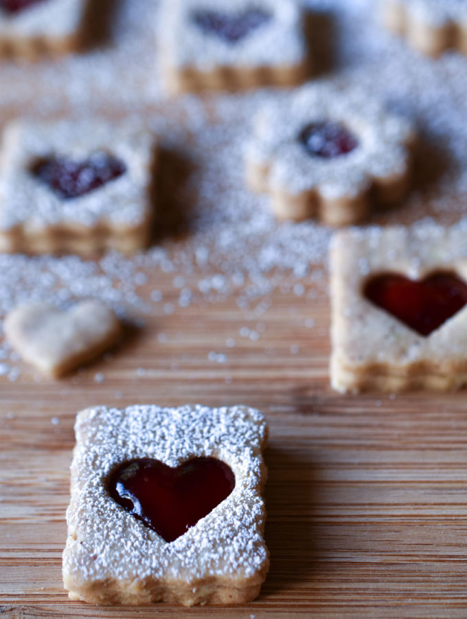 Heart and flower shaped powdered sugar dusted Linzer Cookies on a wood cutting board.