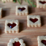 Heart and flower shaped Linzer Cookies on a wood cutting board with green branches and red berries in the background.