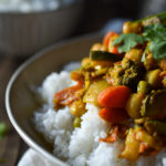 A warm bowl of Takari (Nepalese Vegetable Curry) atop white rice with another bowl of white rice and cup of tea in the background.