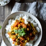 An overview of a warm bowl of Takari (Nepalese Vegetable Curry) atop white rice with a small bowl of white rice a cup of tea and silverware off to the side.