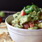 A bowl of Cadillac Guacamole with tortilla chips around and a roma tomato in the background