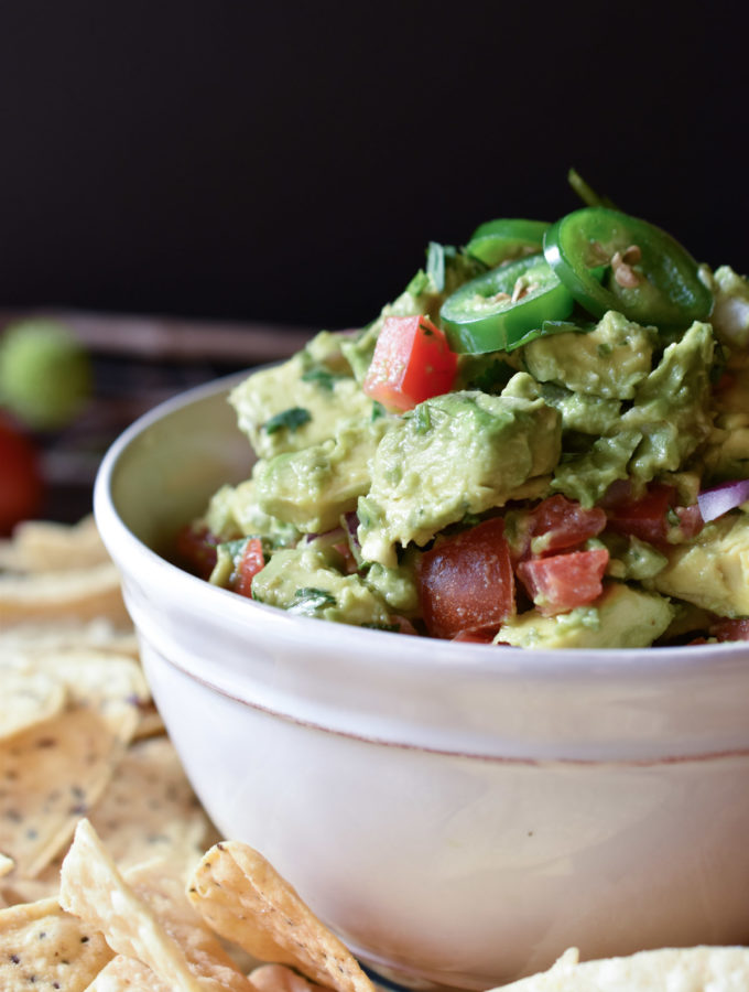 A bowl of Cadillac Guacamole with tortilla chips around and a roma tomato in the background
