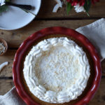 Creamy Cheesecake Pie with Peanut Butter, Honey & Coconut in a decorative napkin with flakes of coconut, a dish of shredded coconut, and a plate with utensils in the background.