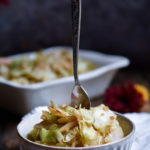 A bowl of Momma's Best Soy Ginger Cabbage with a fork in the middle with a filled serving dish and orange and red flowers in the background.