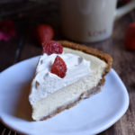 A slice of Classic Cheesecake Pie with Strawberry with another slice, a couple strawberries and a "love" coffee cup in the background.