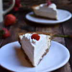 A slice of Classic Cheesecake Pie with Strawberry with another slice, a couple strawberries, twigs and flowers in the background.
