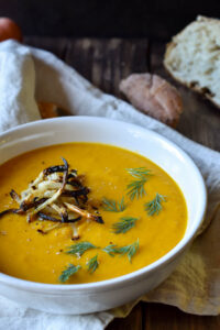 A bowl of Carrot, Orange & Dill Soup with crispy onions a top and bread in the background.