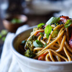 A close up of Easy Udon Noodle Vegetable Stir Fry with a small bowl of green onions and sake in the background