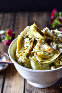 A close up of a bowl of Herb Marinated Artichoke Hearts with vibrant red flowers in the background.