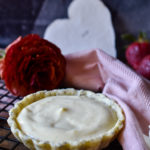 A Vanilla Bean Cream Tart on a rack with a paper heart, pink napkin and flowers in the background.
